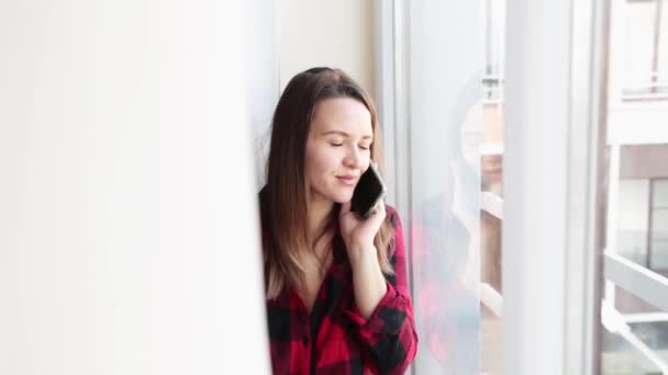 Gelukkige jonge vrouw in een rood geruit shirt gebruikt een mobieltje bij het grote raam. Een ontspannen dame die een bericht typt op haar mobieltje. Vrolijk gelukkig meisje. — Stockvideo