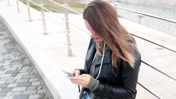 Ung snygg långhårig kvinna bär i trendiga casual kläder sitter ute på gatan och använder sin telefon. söt attraktiv flicka chatta med mobiltelefon. — Stockvideo