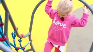 Küçük, aktif, beyaz, sarışın kıvırcık kız oyun parkında spor kompleksine tırmanıyor. Çocuk anaokulunda salıncak yuvasında 