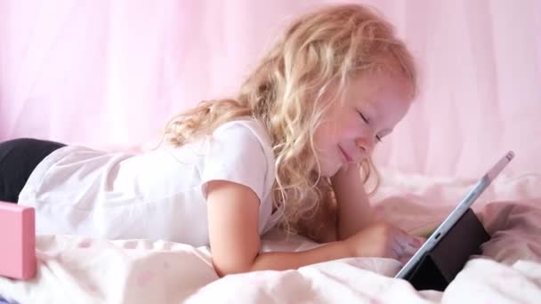 Кавказская любопытная кудрявая милая дошкольница, использующая цифровые планшетные технологии, лежащая в постели одна. Маленький ребенок держит планшет для серфинга интернет-игры дома. Детская техническая зависимость — стоковое видео