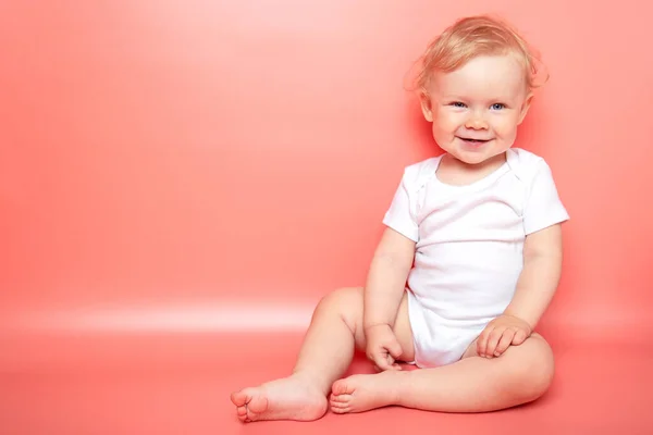 肖像画高加索卷曲金发微笑的女婴穿着白色T恤坐在粉色背景上。复制空间. — 图库照片