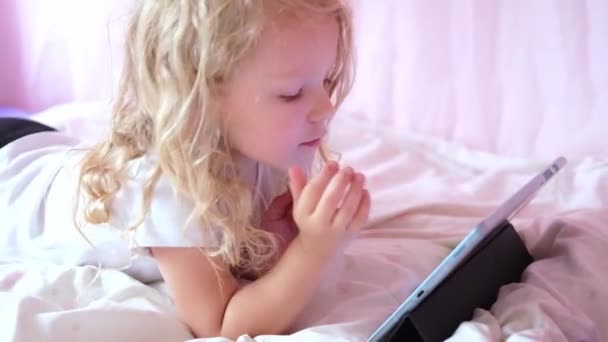 Kaukasische neugierige lockige süße Vorschulkind Mädchen mit digitalen Tablet-Technologie Gerät allein auf dem Bett liegend. Kleines Kind hält Pad Surfen Internet-Spiel zu Hause. Techniksucht bei Kindern — Stockvideo