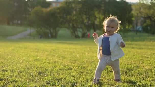 Кавказька мила кучерява дівчинка-дівчинка навчається ходити по зеленій траві в парку на заході сонця. Дитина махає рукою і дивиться на камеру. — стокове відео