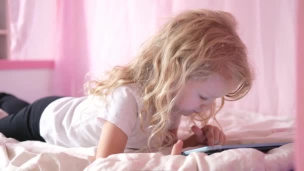 Caucasien curieux frisé mignon préscolaire enfant fille à l'aide de tablette numérique dispositif technologique couché sur le lit seul. Petit enfant tenir pad surf jeu Internet à la maison. Dépendance technologique des enfants — Video