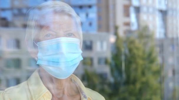 Old woman in protective medical mask through the glass window. Quarantine for coronavirus pandemic. Hope hand and support for recovery from covid-19. elderly people. — Stok video