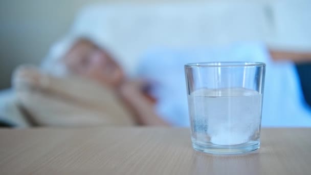 Nahaufnahme Schmerztablette löst sich in Glas auf Tisch im Hintergrund, ältere Frau liegt auf dem Bett. Die alte Frau ist erkältet. Medikamentenprozesskonzept. — Stockvideo