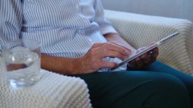 Dijital tablet tutan, kitap okuyan yaşlı kadının ellerini kapat ve evdeki kanepeye otur. Yetişkin bir bayan bilgisayar uygulamaları kullanıyor. Kanepede internet alışverişi yapıyor. alışveriş çevrimiçi kavramı.