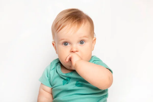 可爱的白种人一岁大的婴儿吸吮大拇指，抓挠他的第一颗牙齿。蹒跚学步的男孩在工作室里拿白色背景开玩笑。幼儿保育和儿童健康概念 图库照片