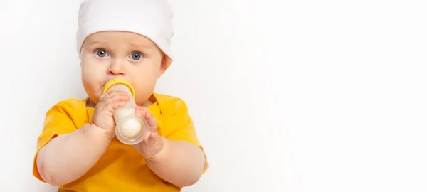 一个可爱的快乐的高加索男婴的肖像，身穿黄色T恤，头戴白色背景的帽子。孩子们从一个有空间的瓶子里喝牛奶。营养和保健概念 免版税图库照片