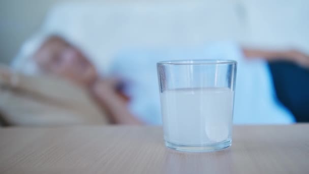 Nahaufnahme Schmerztablette löst sich in Glas auf Tisch im Hintergrund, ältere Frau liegt auf dem Bett. Die alte Frau ist erkältet. Medikamentenprozesskonzept. — Stockvideo