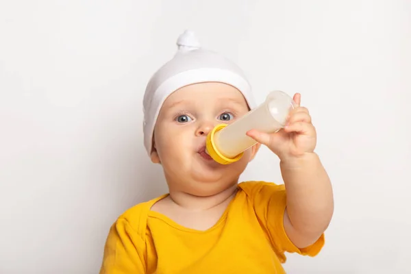 Retrato de un lindo bebé caucásico feliz vestido con una camiseta amarilla y un sombrero sobre fondo blanco. niño bebe leche de un biberón con copyspace. concepto de nutrición y atención sanitaria — Foto de Stock