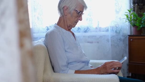 Портрет літньої жінки тримається в руках цифровий планшет читання електронної книги сидить на дивані вдома. старша доросла леді, використовуючи комп'ютерні додатки, які переглядають інтернет-магазини на дивані. стара жінка купує онлайн концепцію — стокове відео