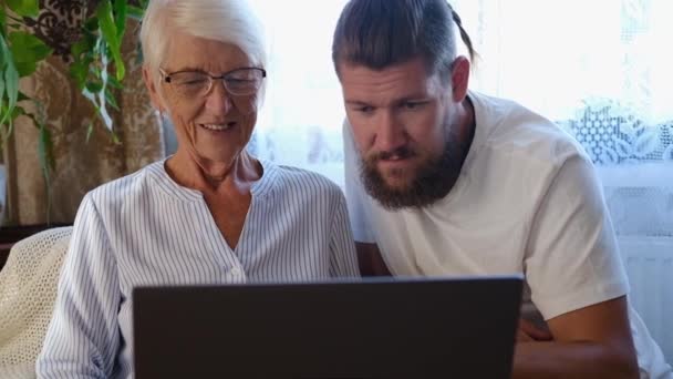 Щаслива старша мати, дорослий син сидить у вітальні на дивані і використовує ноутбук, намагаючись зробити онлайн-платежі. пенсійний спосіб життя, якість сімейного перебування вдома. середнього віку вдалося допомогти мамі — стокове відео