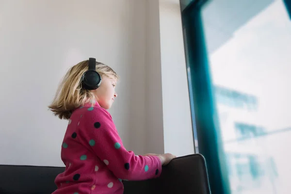 Kleines Mädchen mit Kopfhörern, das zu Hause aus dem Fenster schaut — Stockfoto