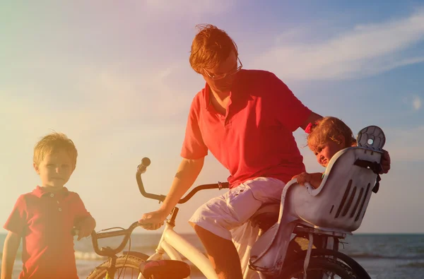 Père avec deux enfants faisant du vélo au coucher du soleil mer — Photo