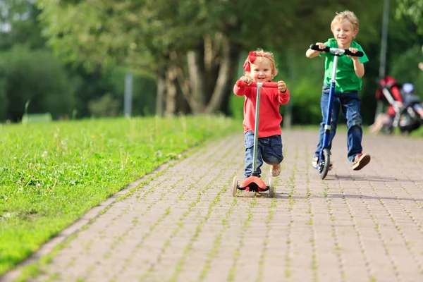 Kleiner Junge und kleines Mädchen fahren Roller im Sommerpark — Stockfoto