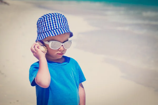 Μικρό αγόρι ακρόασης θαλασσινών κοχυλιών στην παραλία — Φωτογραφία Αρχείου