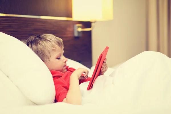Pequeño niño mirando almohadilla táctil acostado en la cama de la habitación de hotel —  Fotos de Stock