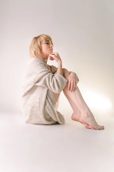 Blonde young woman dressed in large white cashmere sweater and seating on white whole-floor — Stock Photo, Image