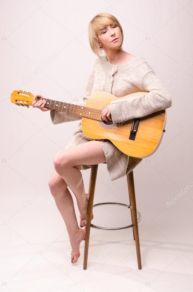 Young blonde lady in gray sweater sitting on chair and playing acoustic guitar