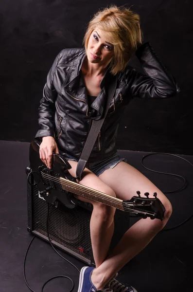 Beautiful young blonde dressed in black leather jacket with electric guitar on a black background — Stock Photo, Image