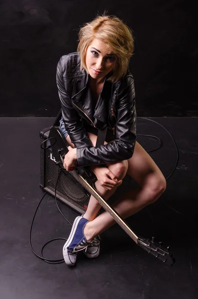 Beautiful young blonde dressed in black leather jacket with electric guitar on a black background — Stock Photo, Image