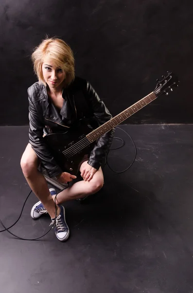 Beautiful young blonde dressed in black leather jacket with electric guitar on a black background — Stock Photo, Image