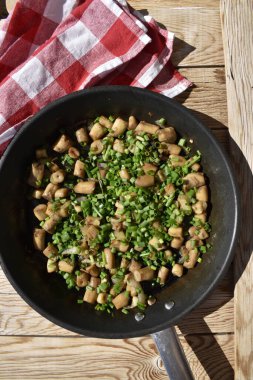 Fried mushroom stems sprinkled with green onions in a pan on a wooden background with a red checkered kitchen napkin, top view
