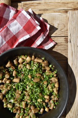 Fried mushroom stems sprinkled with green onions in a pan on a wooden background with a red checkered kitchen napkin, top view