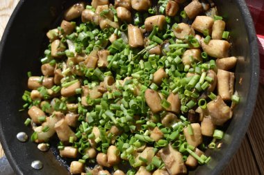 Fried mushroom stems sprinkled with green onions in a pan on a wooden background with a red checkered kitchen napkin, top view