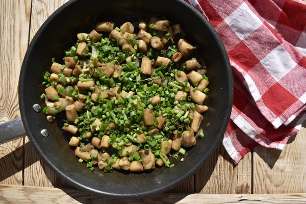 Fried mushroom stems sprinkled with green onions in a pan on a wooden background with a red checkered kitchen napkin, top view