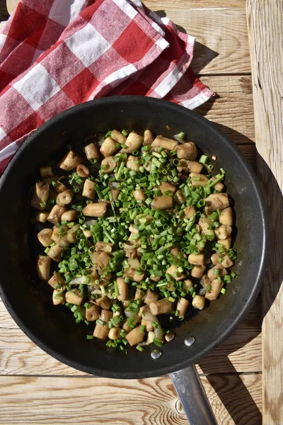 Fried mushroom stems sprinkled with green onions in a pan on a wooden background with a red checkered kitchen napkin, top view