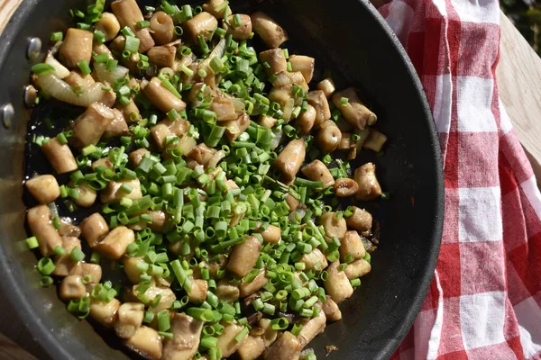 Fried mushroom stems sprinkled with green onions in a pan on a wooden background with a red checkered kitchen napkin, top view