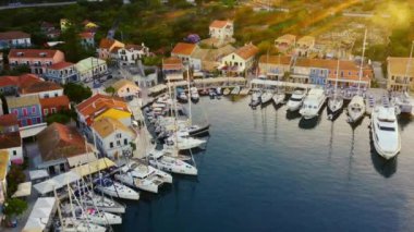 İnsansız hava aracı Yunanistan 'daki güzel marina ve Fiskardo kasabasının üzerinde geriye doğru uçuyor. Yat limanında bir sürü tekne var. Günbatımında güzel bir akşam, çerçevenin sağ tarafında güneş parlıyor. Arka planda dağlar var. 1080p