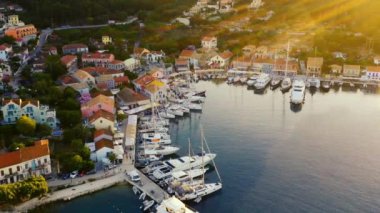 İnsansız hava aracı Yunanistan 'daki güzel marina ve Fiskardo kasabasının üzerinde uçuyor. Yat limanında bir sürü tekne var. Günbatımında güzel bir akşam, çerçevenin sağ tarafında güneş parlıyor. Arka planda dağlar var. 4K