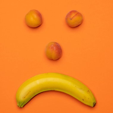 Olgun sarı muz ve taze şeftalilerin renkli arka planda somurtkan bir yüz oluşturduğu bir fotoğraf.