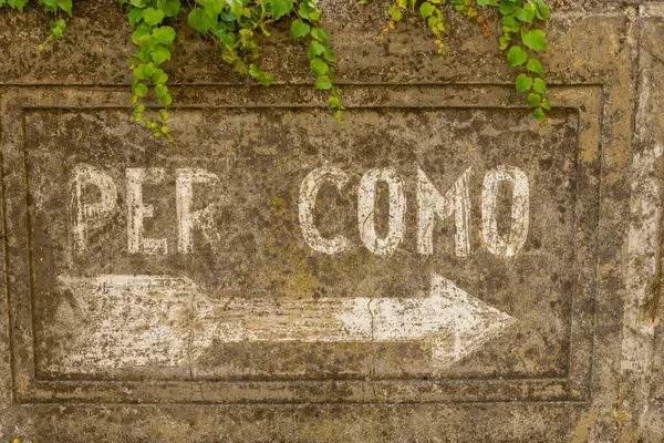 Old painted sign on a wall, showing direction to the city of Como, Lombardy, Italy. A lot of the white paint has fallen of the old stone wall where it has been painted. Green plants are hanging above the sign.