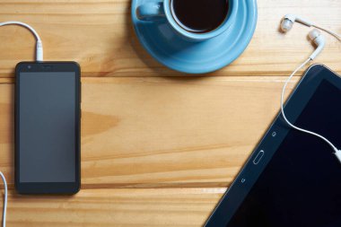 Üzerinde siyah cep telefonu, tablet, kulaklık ve bir fincan kahve olan ahşap masa manzarası. Çalışma alanı kavramı, ev ofisi, çalışma ya da dinlenme yeri. Merkezde boşluk kopyala.
