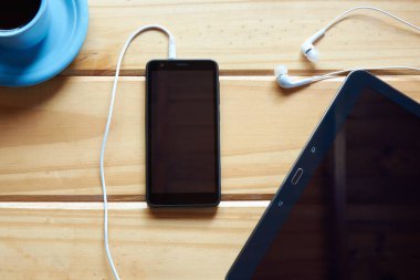 Üzerinde siyah cep telefonu, tablet, kulaklık ve bir fincan kahve olan ahşap masa manzarası. Çalışma alanı kavramı, ev ofisi, çalışma ya da dinlenme yeri. Merkezde boşluk kopyala.