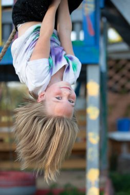 Güzel küçük kız oyun bahçesinde baş aşağı asılı duruyor. Küçük kız kameraya gülümsüyor..