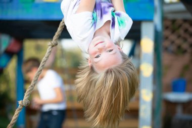 Güzel küçük kız oyun bahçesinde baş aşağı asılı duruyor. Küçük kız kameraya gülümsüyor..