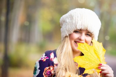 Akçaağaç yaprağı olan sıcak şapkalı güzel bir kız kameraya bakar ve gülümser..