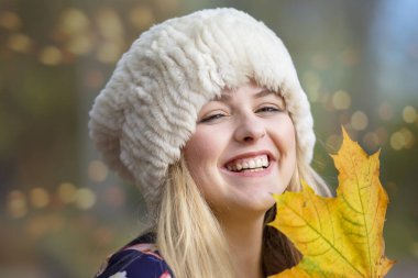 Akçaağaç yaprağı olan sıcak şapkalı güzel bir kız kameraya bakar ve gülümser..