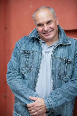 Kot ceketli yaşlı bir adam kameraya bakar ve gülümser. 50 yaşında, dik kafalı, beyaz saçlı, iri yapılı bir adam..