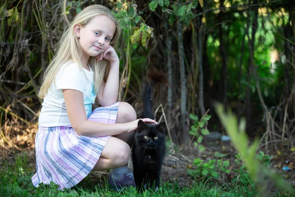 金发小女孩在绿树成荫的背景上和一只黑猫玩耍 — 图库照片