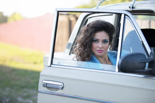 Funny young woman in a denim jacket with make-up in the style of the eighties looks through the car window.