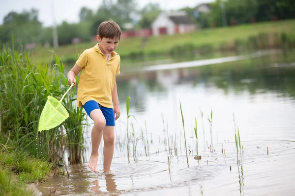 A little boy with a fishing net walks near the lake and looks at the water.