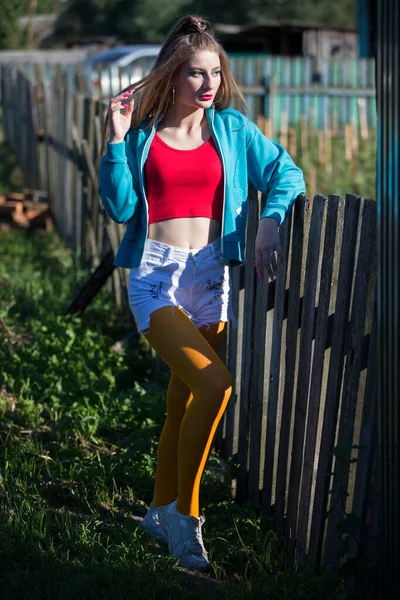 A beautiful girl in bright clothes stands near a wooden fence. Woman in the style of the 90s.