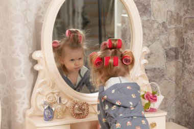 İki yaşında bir kız. Bigudili küçük bir kız makyaj masasındaki aynaya bakıyor. Parti hazırlığı. Görünüşünüzden memnun kalmadınız..