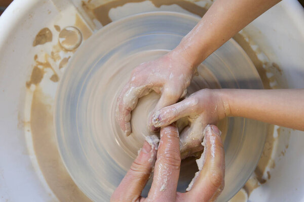 Крупный план рук гончара и руки ребенка с предметом на гончарном круге. Работа с глиной. Мастерская глины. Обучение ремеслу.